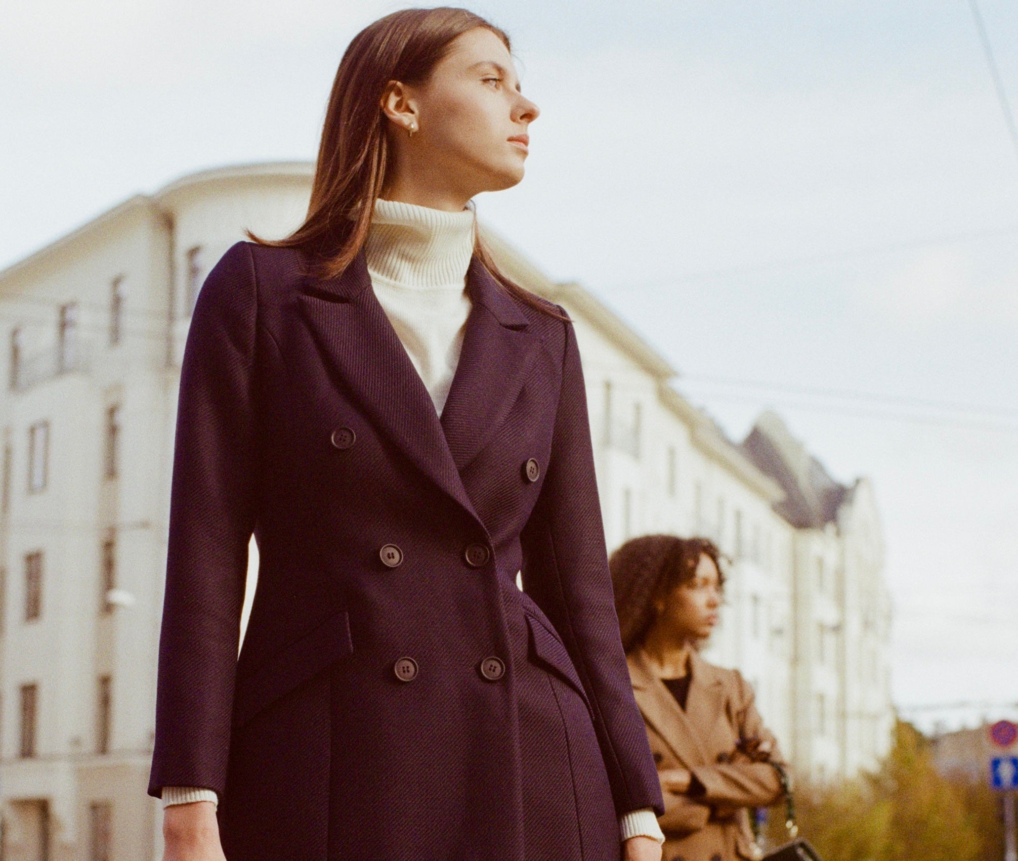 Woman in a double-breasted jacket standing on a city street with another person in the background wearing double-breasted coat.